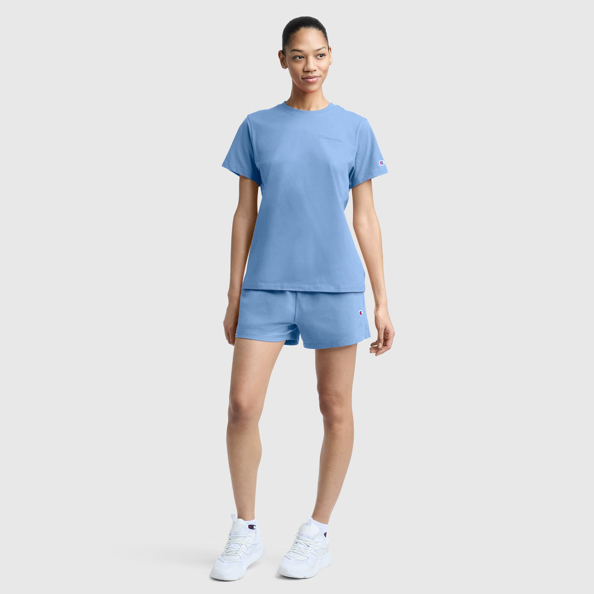 A woman stands against a plain background, wearing the Champion Practice Short in light blue paired with a matching T-shirt and white sneakers. Her hair is pulled back, and she smiles slightly with one hand by her side and the other relaxed.