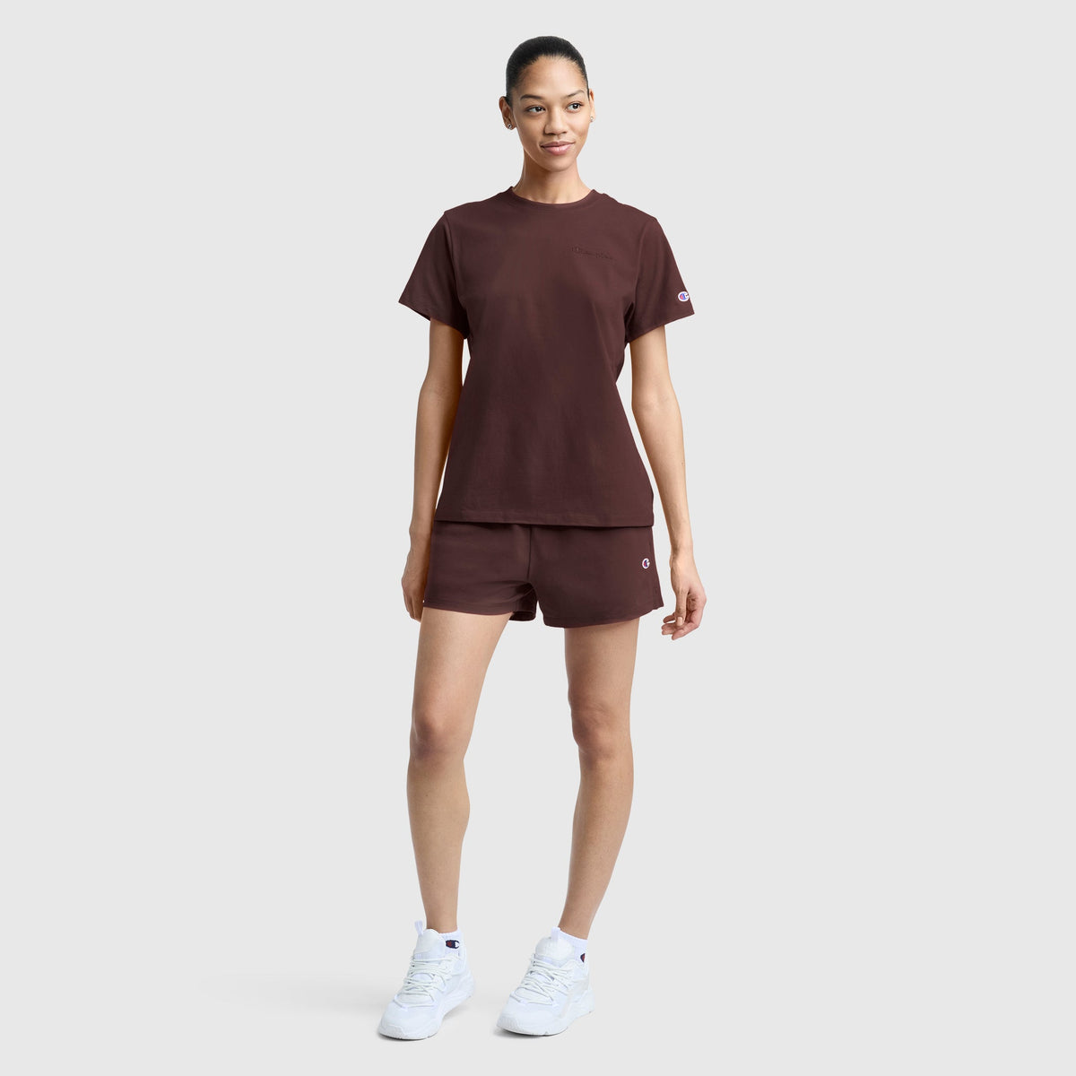 A woman stands against a plain background, wearing the Champion Practice Short in a matching maroon set with a t-shirt and white sneakers. Her hair is tied back, and she looks ahead with a neutral expression.