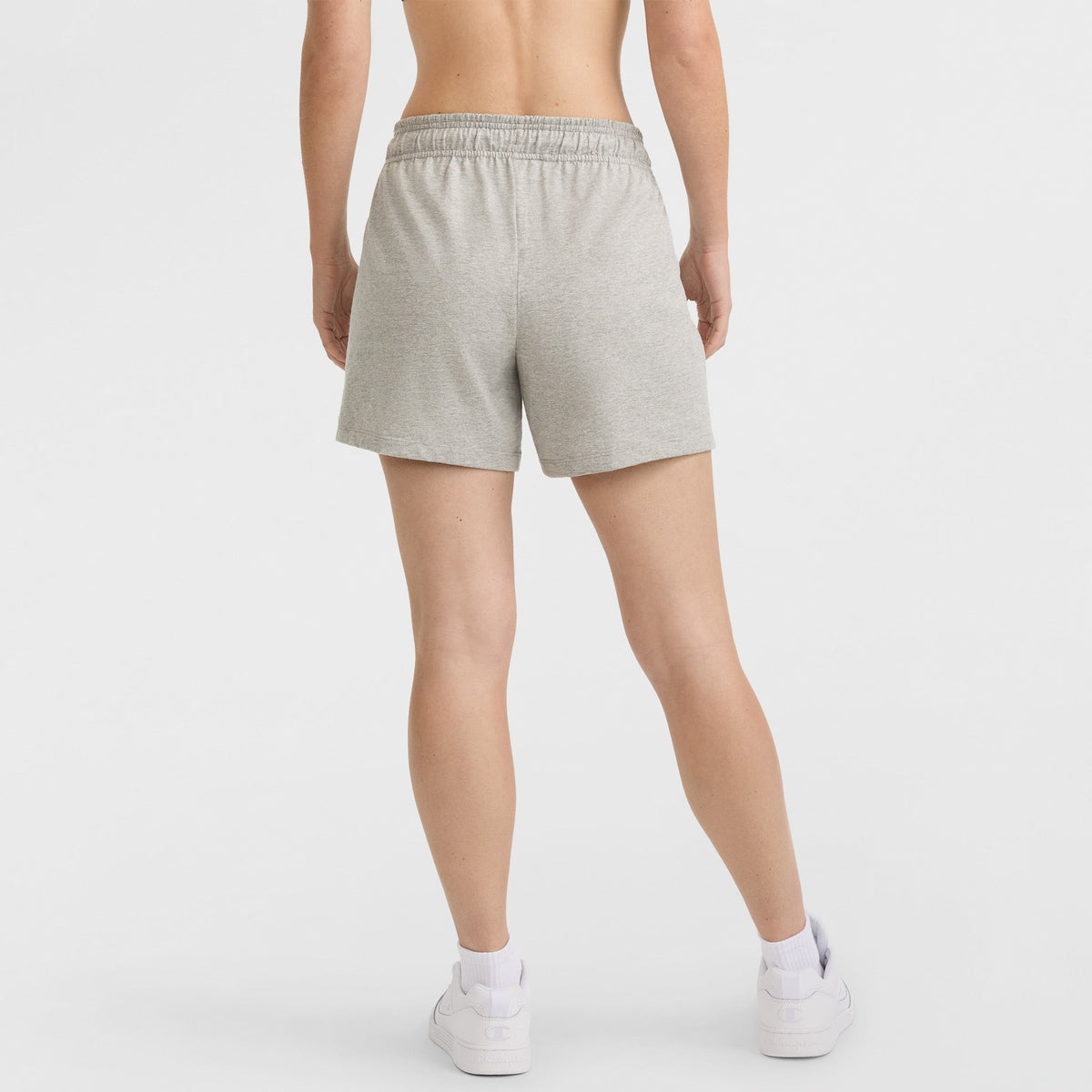A person stands facing away, wearing Champion 4" Lightweight Lounge Shorts with a C Logo in light gray, paired with white socks and white sneakers. The background is plain white.