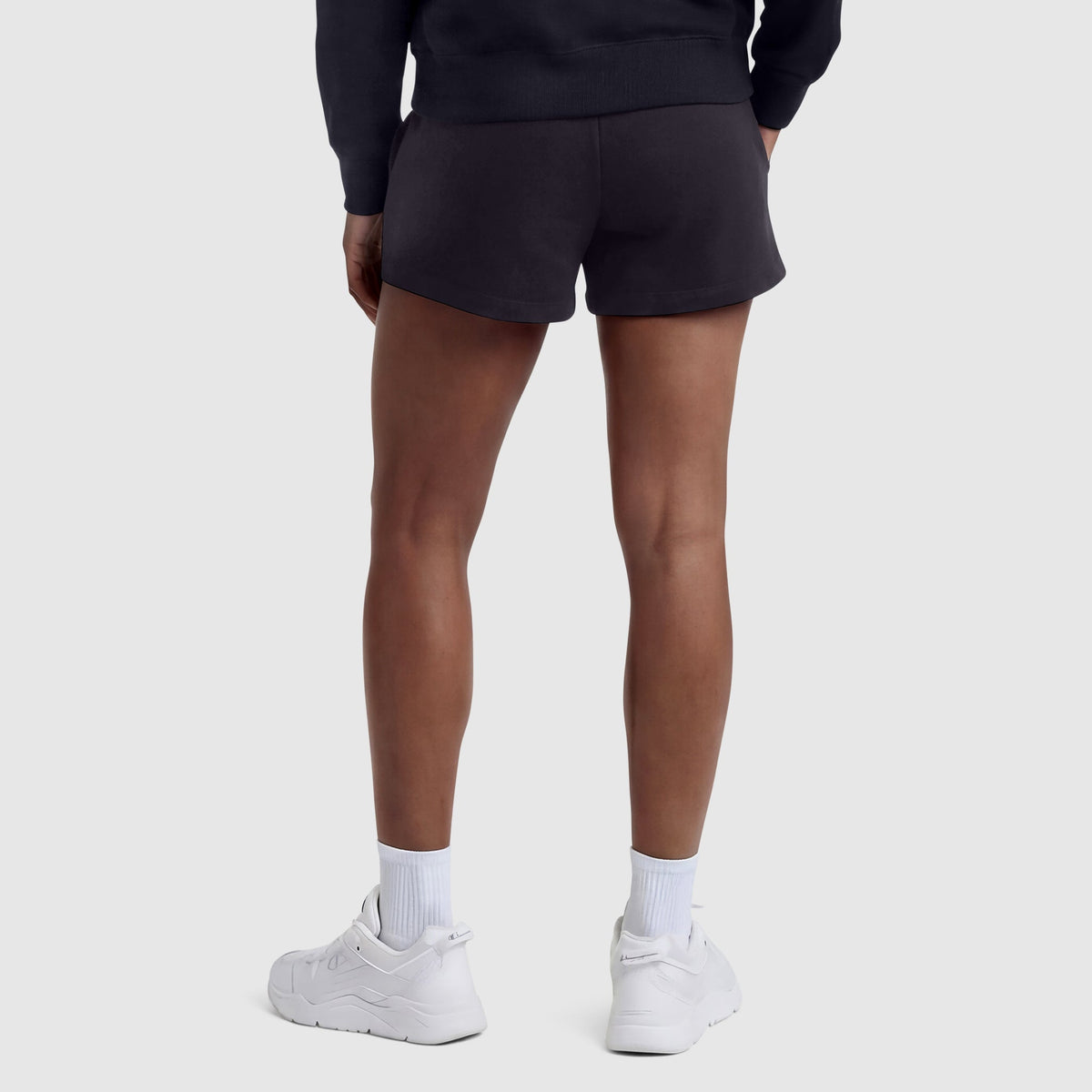 A person stands with their back to the camera, wearing a black sweatshirt and Champion 4" Lightweight Lounge Shorts with the C Logo—crafted from lightweight cotton fabric and designed with an adjustable waistband—paired with white crew socks and white athletic shoes against a plain light background.
