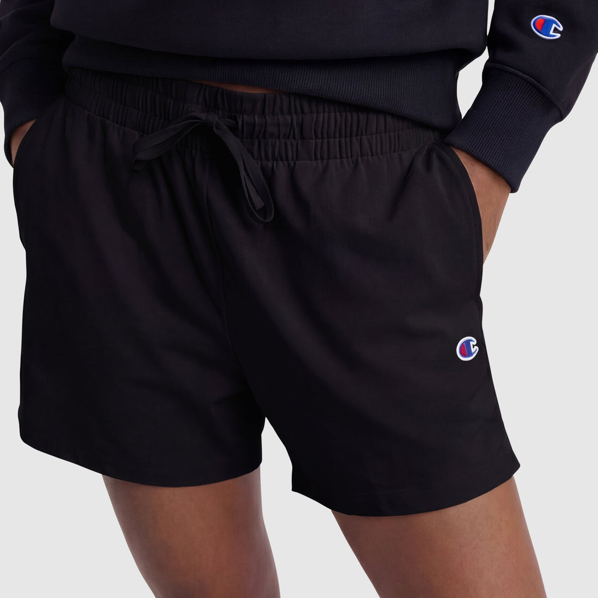 A person is wearing black Champion 4" Lightweight Lounge Shorts, C Logo, crafted from lightweight cotton with an adjustable waistband, paired with a matching black Champion sweatshirt. Hands are in pockets, with visible logo patches on both pieces, standing against a plain light background.