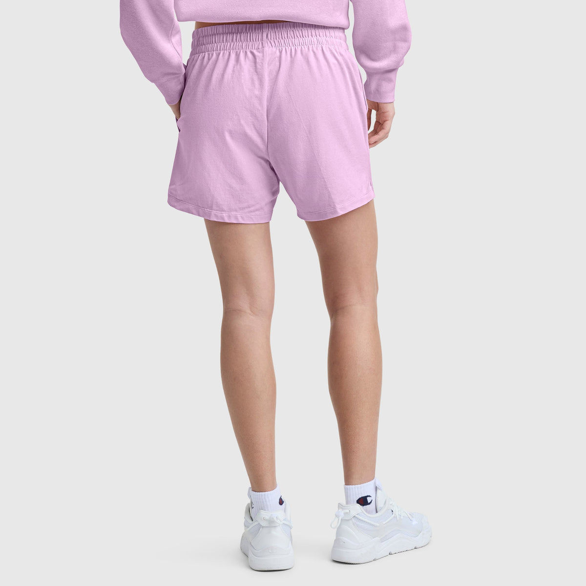 A person wearing Champion 4″ Lightweight Lounge Shorts, C Logo, in light purple along with a matching sweatshirt, white socks, and white sneakers stands on a light gray background with their back to the camera.