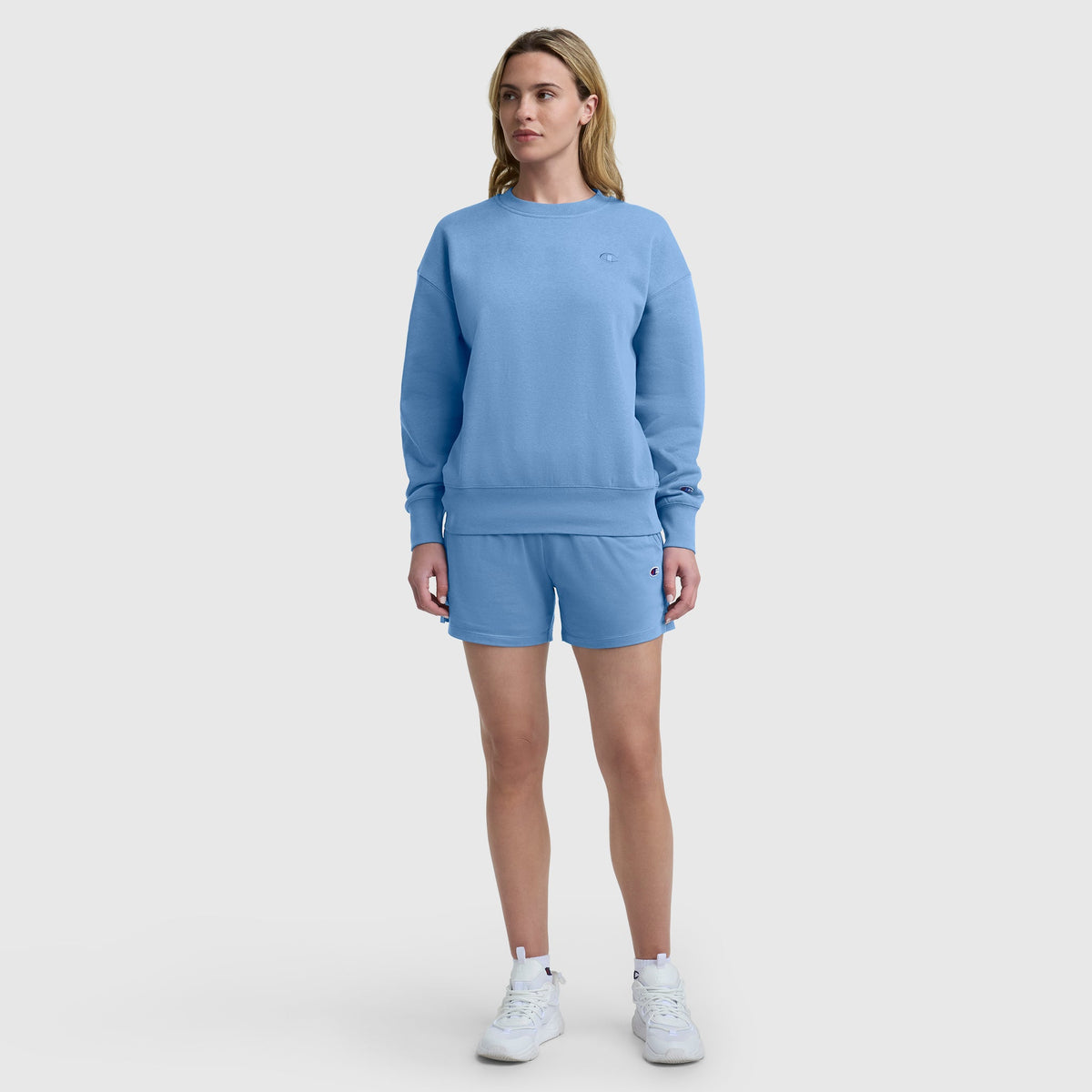 A person stands facing forward against a plain white background, wearing a light blue sweatshirt, Champion 4″ Lightweight Lounge Shorts with the C Logo featuring an elastic waistband, and white sneakers.