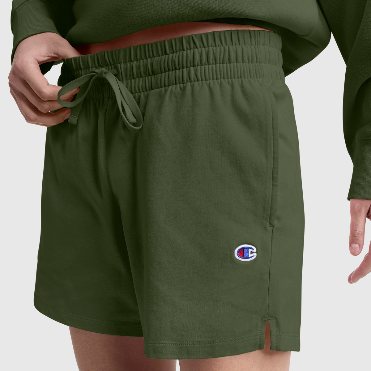 A person wearing Champion 4" Lightweight Lounge Shorts, featuring an olive green color, a drawstring waist, and a small C logo on the left leg, shown from the waist down. These shorts are made from a cotton blend for everyday lightweight comfort.