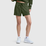 A person stands against a plain light background, showing only their lower body, wearing Champion’s 4" Lightweight Lounge Shorts with C Logo in olive green, paired with a matching green sweatshirt, white Champion sneakers, and white socks.