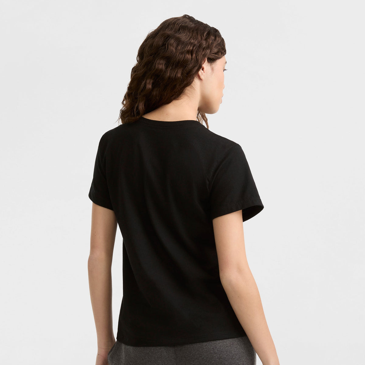 A person with wavy brown hair stands facing away, wearing the Champion Classic T-Shirt with tonal left chest script in black and dark gray pants, set against a light gray background.