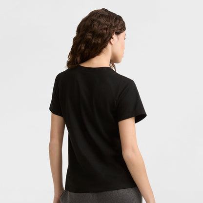 A person with wavy brown hair stands facing away, wearing the Champion Classic T-Shirt with tonal left chest script in black and dark gray pants, set against a light gray background.