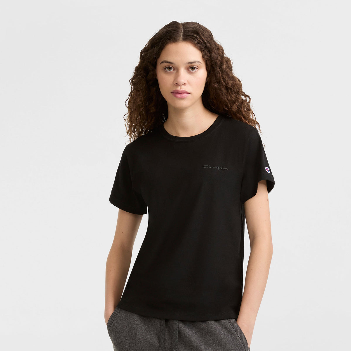 A young woman with long curly hair wears a black Champion Classic T-Shirt, Tonal Left Chest Script, and gray pants, standing against a white background with hands in her pockets, looking directly at the camera.