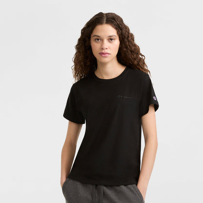 A young woman with long curly hair wears a black Champion Classic T-Shirt, Tonal Left Chest Script, and gray pants, standing against a white background with hands in her pockets, looking directly at the camera.