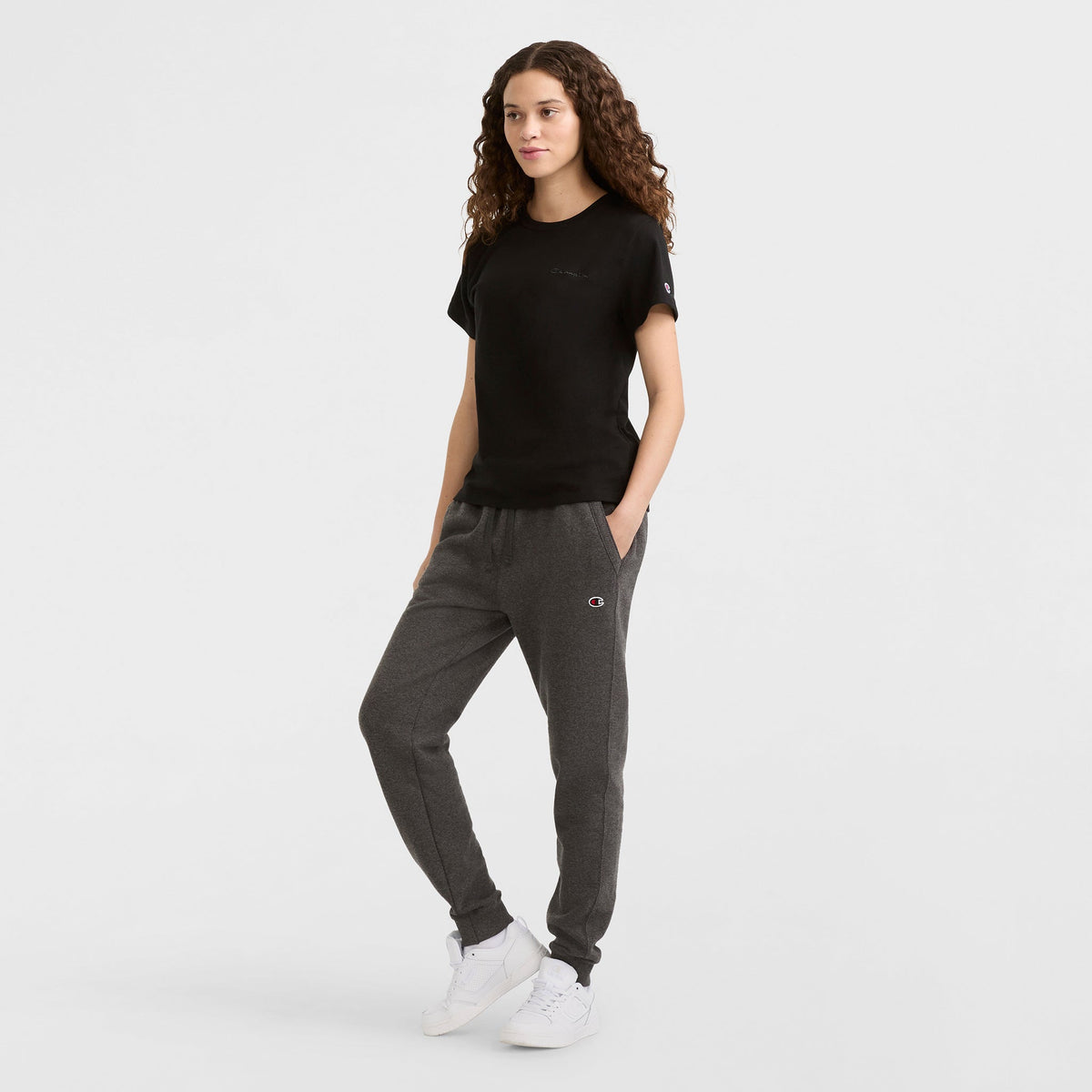 A woman with long curly hair models the Champion Classic T-Shirt, Tonal Left Chest Script. She pairs it with gray sweatpants and white sneakers, standing against a plain white background with her hands in her pockets.