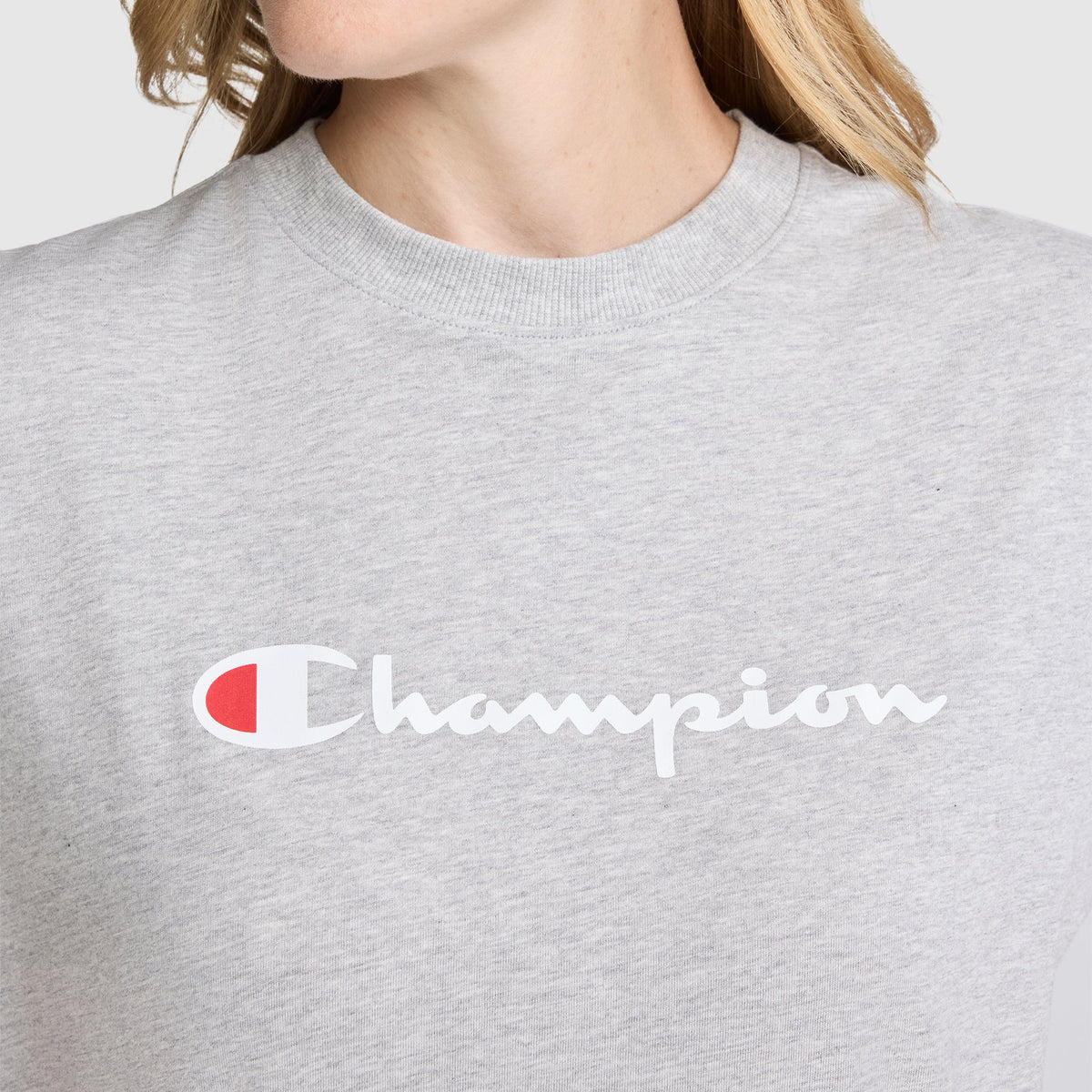 A person wearing a light gray Champion Classic T-Shirt, Script Logo, featuring the white and red script logo across the chest. The person's face is partially out of frame.