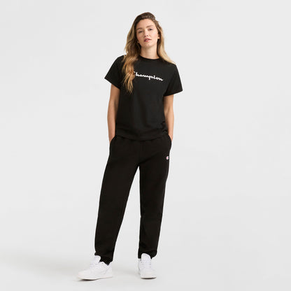 A person with long, wavy hair stands against a plain white background wearing the Champion Classic T-Shirt with script logo, black sweatpants, and white sneakers. Their hands are in their pockets.