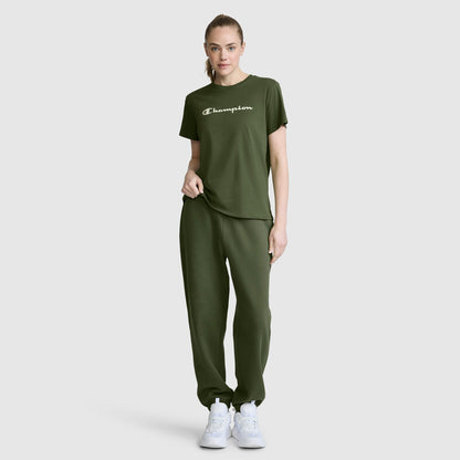 A woman stands against a plain light gray background wearing an olive green Champion Classic T-Shirt with the script logo and matching sweatpants, paired with white sneakers.
