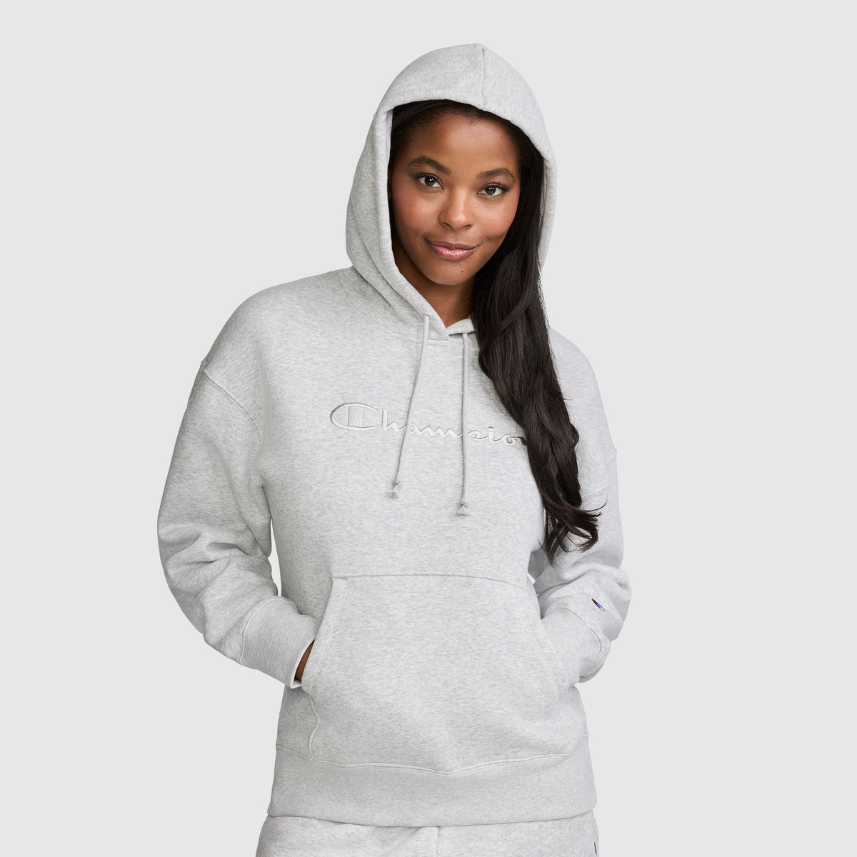 A woman wearing a light gray Champion Tonal Script Freshman Powerblend Hoodie with "Champion" embroidered on the front stands with her hands in the front pocket, smiling slightly against a plain, light background. This cozy hoodie is crafted from soft fleece, providing both comfort and style.