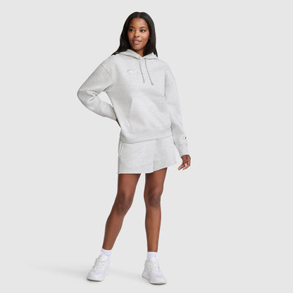 A woman stands in a Champion Tonal Script Freshman Powerblend Hoodie and matching shorts with white socks and sneakers, smiling with one hand on her hip against a plain light background.