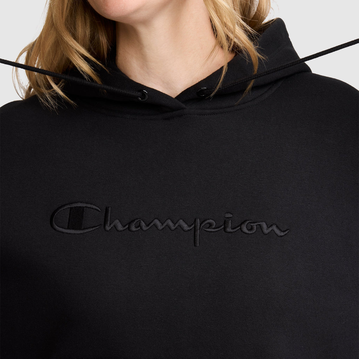A person wears a black Champion Tonal Script Freshman Powerblend Hoodie, with "Champion" embroidered in black across the chest; only the lower face, neck, and some hair are visible.