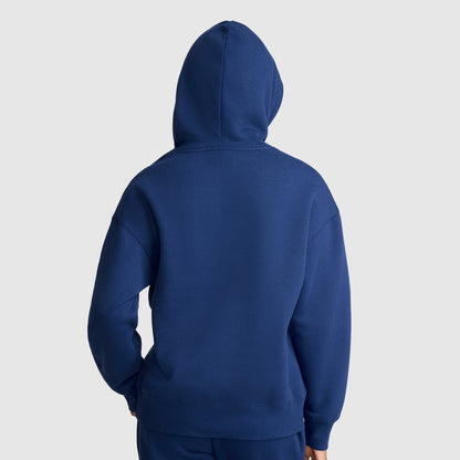A person wearing a Champion Tonal Script Freshman Powerblend Hoodie, crafted from midweight fleece, stands with their back facing the camera, hands resting in the front pocket, against a plain light gray background.