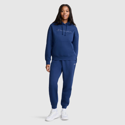 A woman wearing a matching navy blue Champion Tonal Script Freshman Powerblend Hoodie and joggers set, crafted from soft midweight fleece, with white sneakers stands facing forward against a light gray background.