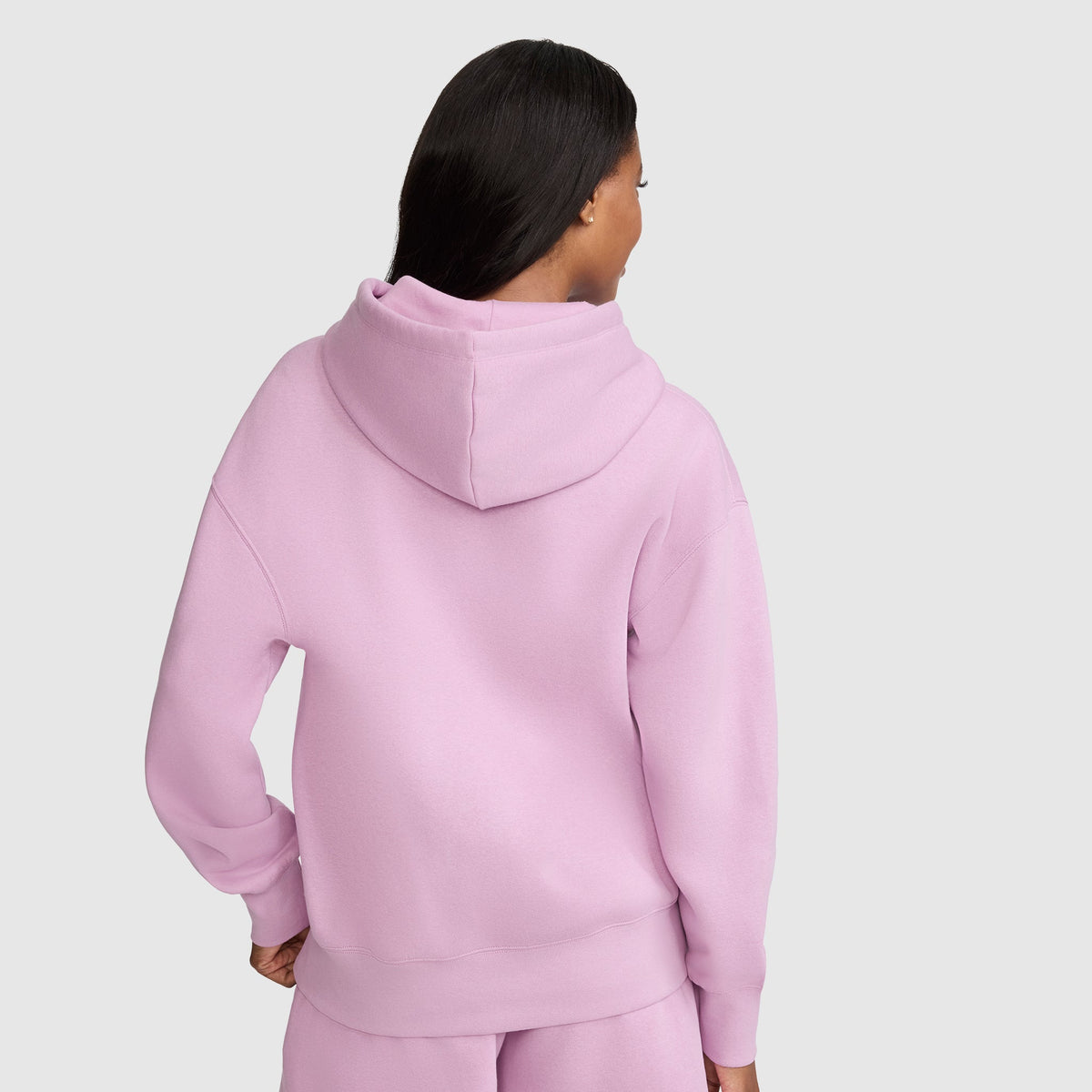 A person with long dark hair wears a light pink Champion Tonal Script Freshman Powerblend Hoodie and matching sweatpants, standing with their back facing the camera against a plain light gray background.