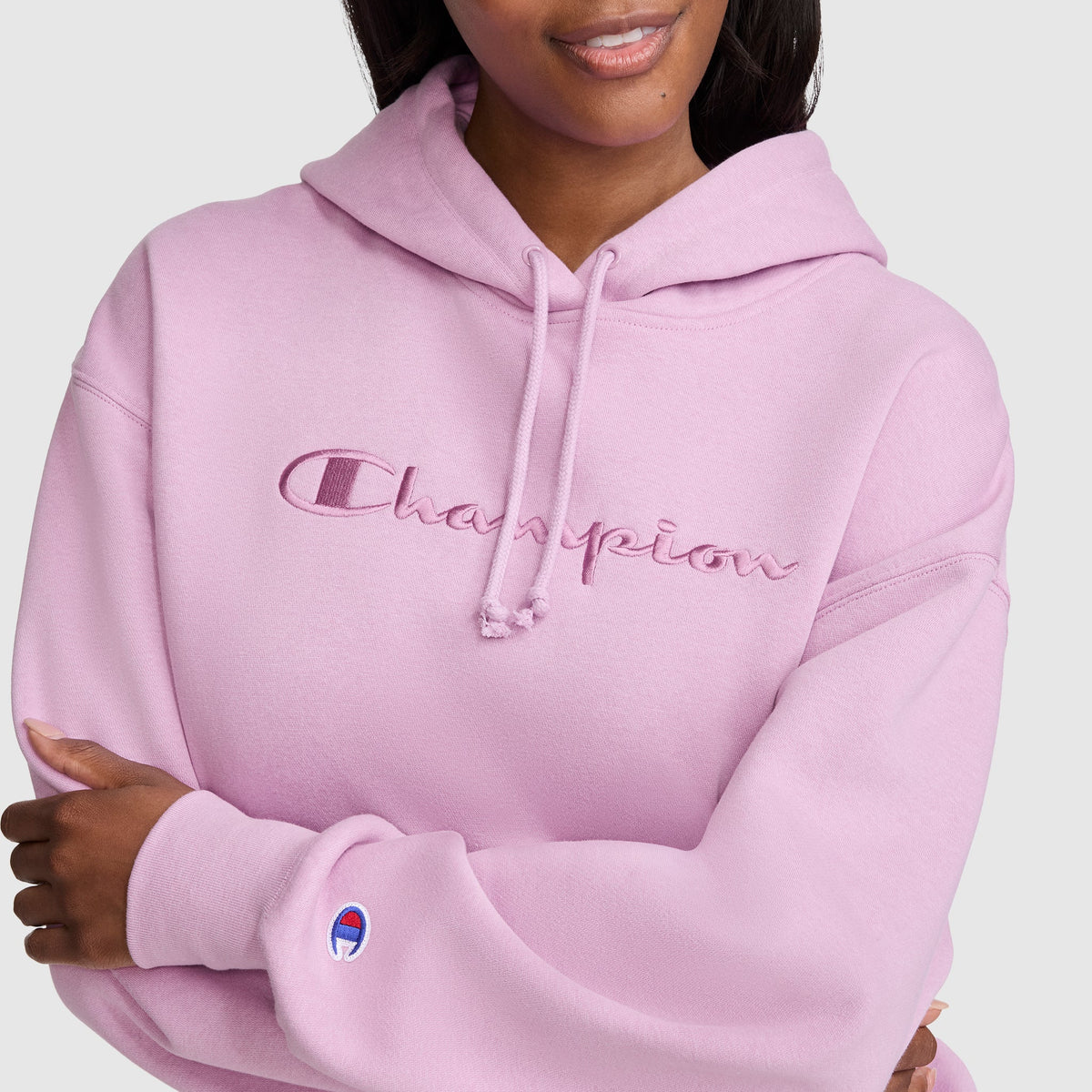 A woman wears a light pink Champion Tonal Script Freshman Powerblend Hoodie, featuring “Champion” embroidered across the front and the brand’s logo on the sleeve. This cozy fleece hoodie is styled as a women’s sweatshirt; she crosses her arms and smiles slightly.
