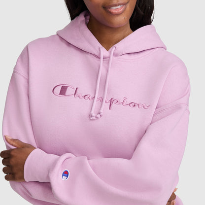 A woman wears a light pink Champion Tonal Script Freshman Powerblend Hoodie, featuring “Champion” embroidered across the front and the brand’s logo on the sleeve. This cozy fleece hoodie is styled as a women’s sweatshirt; she crosses her arms and smiles slightly.