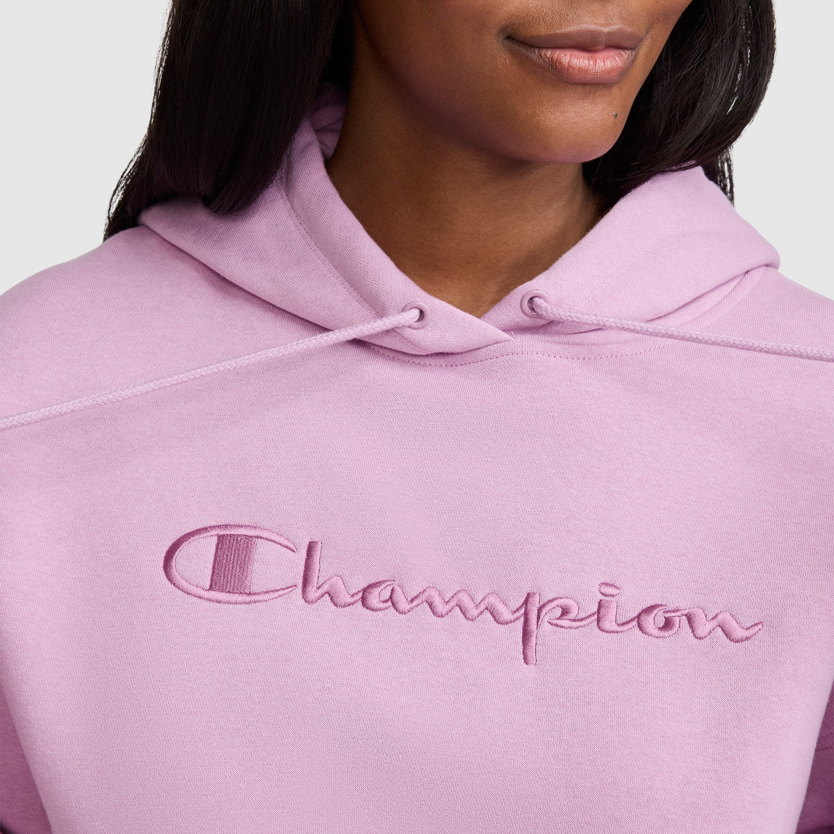 A person wearing a light pink Champion Tonal Script Freshman Powerblend Hoodie, featuring the brand name “Champion” embroidered in matching pink across the chest. Only the lower face and upper torso are visible, showcasing this cozy women’s sweatshirt.