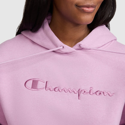 A person wearing a light pink Champion Tonal Script Freshman Powerblend Hoodie, featuring the brand name “Champion” embroidered in matching pink across the chest. Only the lower face and upper torso are visible, showcasing this cozy women’s sweatshirt.