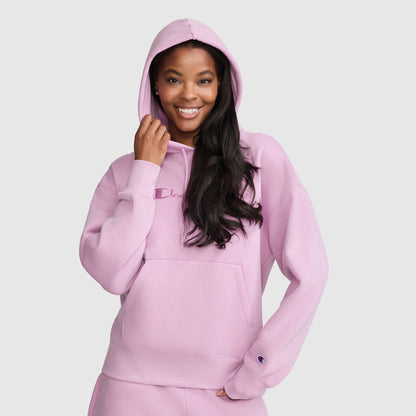 A woman smiles while wearing the Champion Tonal Script Freshman Powerblend Hoodie in light pink, featuring a matching hood and a front pocket, standing against a plain light background. This cozy women’s sweatshirt from Champion is perfect for casual comfort and effortless style.
