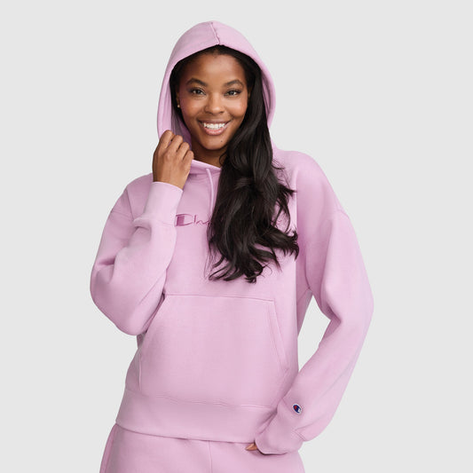 A woman smiles while wearing the Champion Tonal Script Freshman Powerblend Hoodie in light pink, featuring a matching hood and a front pocket, standing against a plain light background. This cozy women’s sweatshirt from Champion is perfect for casual comfort and effortless style.