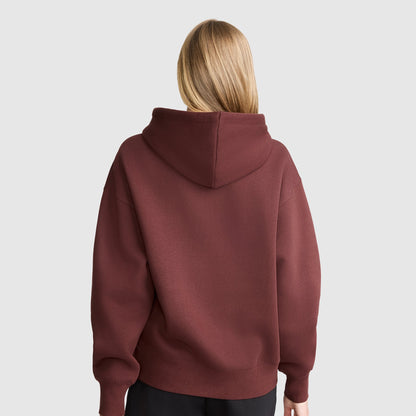 A person with straight, blonde hair is standing with their back to the camera, wearing a maroon Champion Tonal Script Freshman Powerblend Hoodie and black pants against a plain, light gray background.