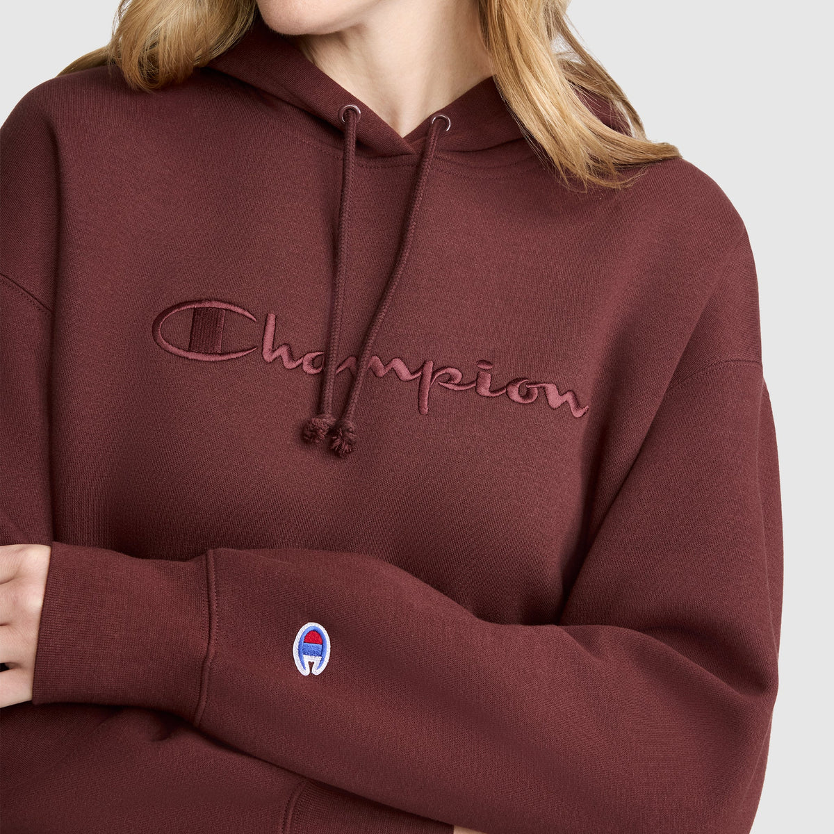 A person wears the Champion Tonal Script Freshman Powerblend Hoodie in maroon, featuring soft fleece fabric, an embroidered tonal script logo on the chest, and a small Champion emblem on the sleeve. Only their lower face, neck, and upper body are visible.