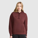 A woman stands against a plain light gray background, wearing the Champion Tonal Script Freshman Powerblend Hoodie in maroon—crafted from cozy fleece with a front pocket—and paired with black pants.