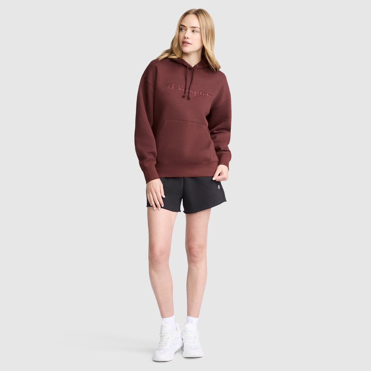 A woman stands wearing the Champion Tonal Script Freshman Powerblend Hoodie in maroon, paired with black shorts, white socks, and white sneakers against a plain light gray background.