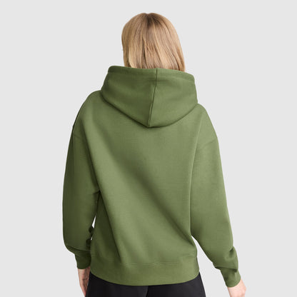 A person with blonde hair is shown from the back, wearing a Champion Tonal Script Freshman Powerblend Hoodie in olive green and black pants, standing against a plain light gray background.