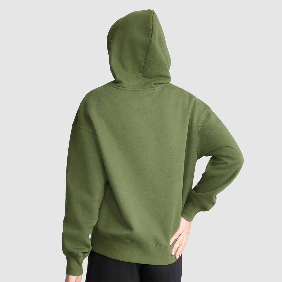 A person wearing a green Champion Tonal Script Freshman Powerblend Hoodie stands with their back to the camera, left hand by their side and right hand resting on their hip, against a plain light gray background.