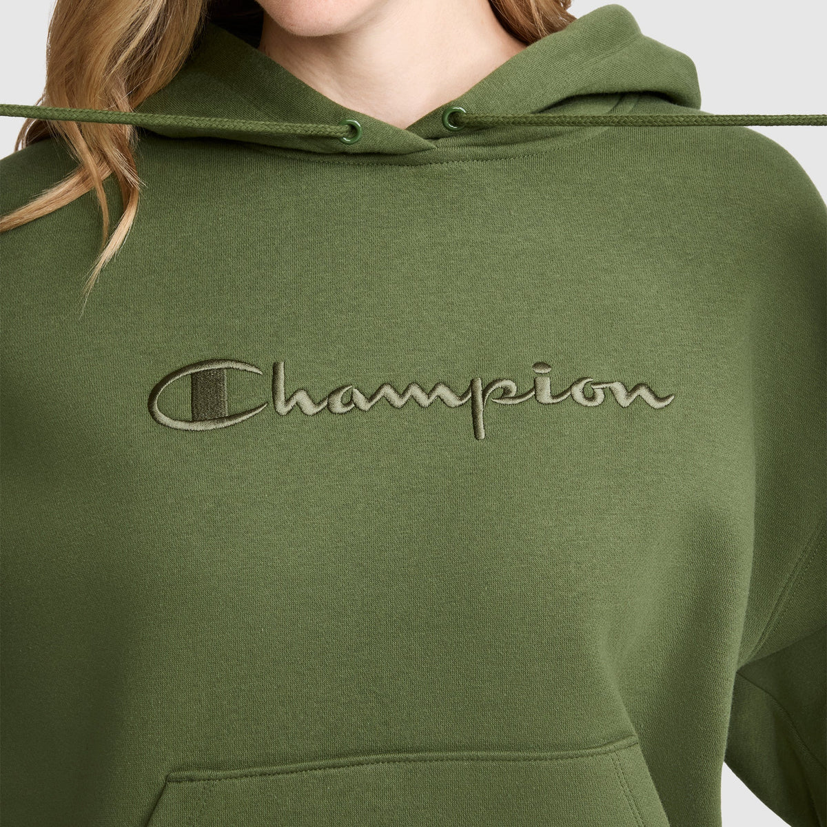 A person wearing a Tonal Script Freshman Powerblend Hoodie by Champion in olive green, featuring the brand's tonal script logo embroidered across the chest. Only their lower face and upper torso are visible, showcasing the classic style of this women's Champion sweatshirt.