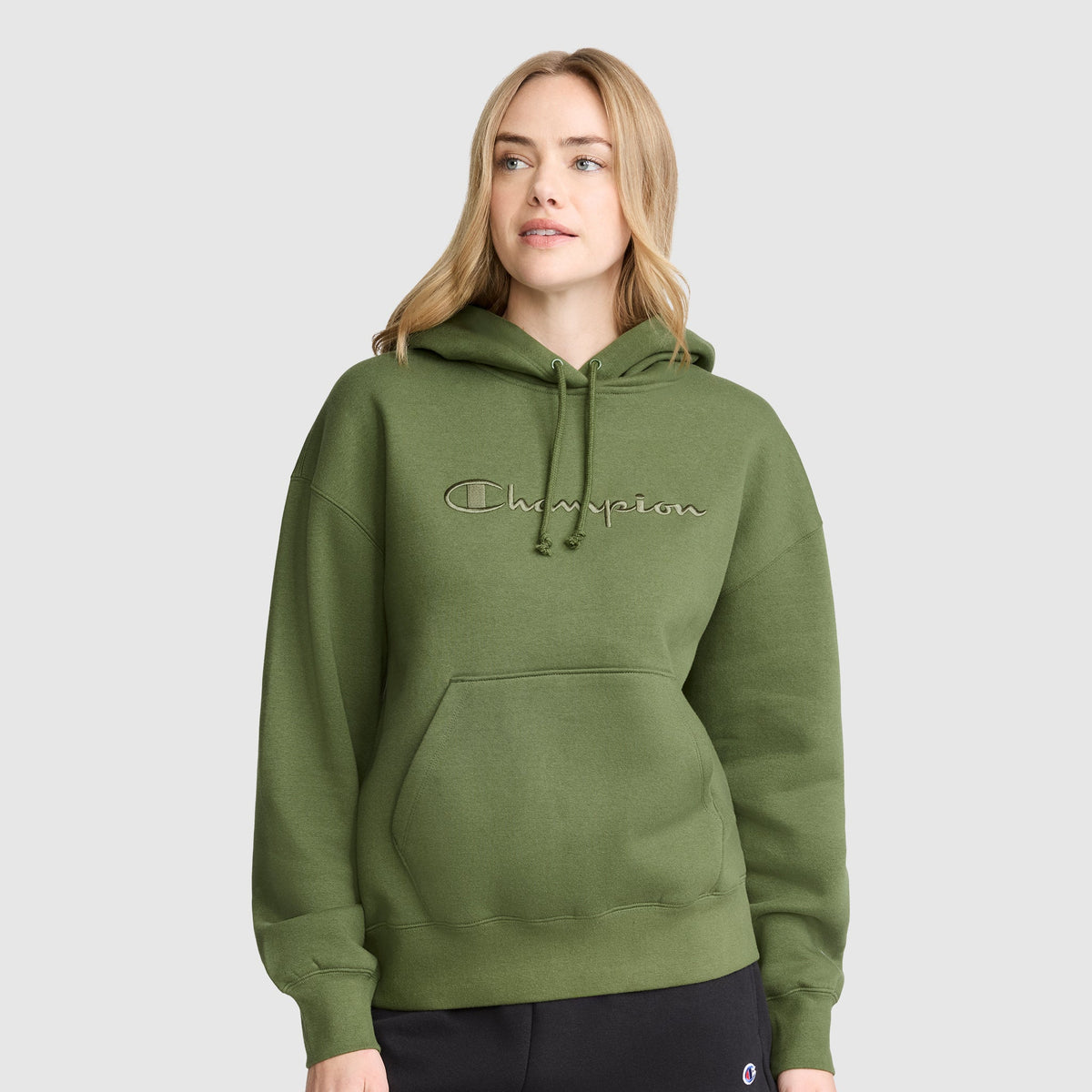 A woman wearing a Champion Tonal Script Freshman Powerblend Hoodie in olive green, featuring a front pocket, stands against a plain gray background, showcasing the comfortable style of this women’s hooded sweatshirt.
