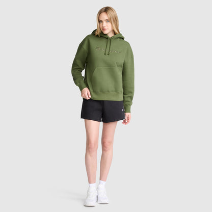 A woman stands wearing the Champion Tonal Script Freshman Powerblend Hoodie in green, featuring the classic Champion text, paired with black shorts, white socks, and white sneakers against a plain light gray background.