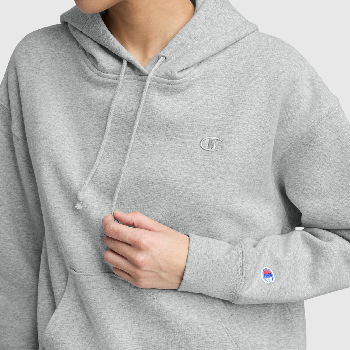Someone wears a light grey Champion Small C Freshman Powerblend Hoodie, made of midweight fleece with an embroidered logo on the chest, a brand patch on the sleeve, an adjustable drawcord hood, and one hand touching the front pocket.