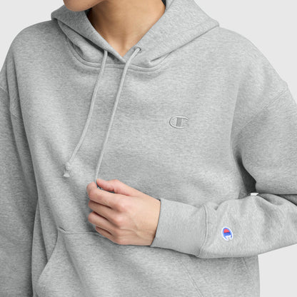 Someone wears a light grey Champion Small C Freshman Powerblend Hoodie, made of midweight fleece with an embroidered logo on the chest, a brand patch on the sleeve, an adjustable drawcord hood, and one hand touching the front pocket.