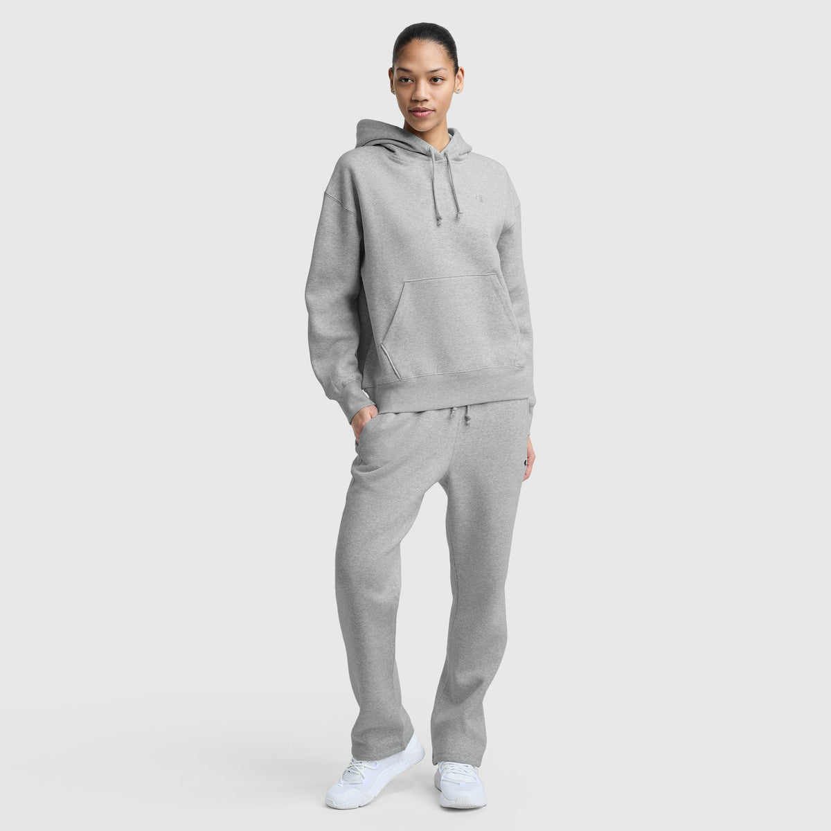 A person stands facing forward in a light gray Champion Small C Freshman Powerblend Hoodie and matching sweatpants with an adjustable drawcord hood and white sneakers, against a plain light gray background.