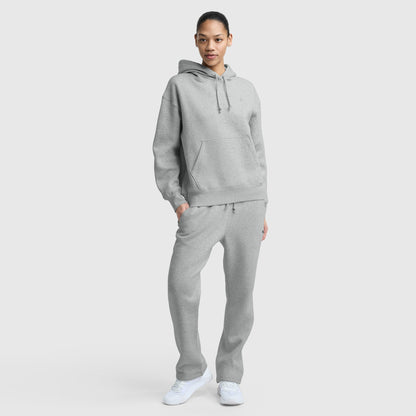 A person stands facing forward in a light gray Champion Small C Freshman Powerblend Hoodie and matching sweatpants with an adjustable drawcord hood and white sneakers, against a plain light gray background.