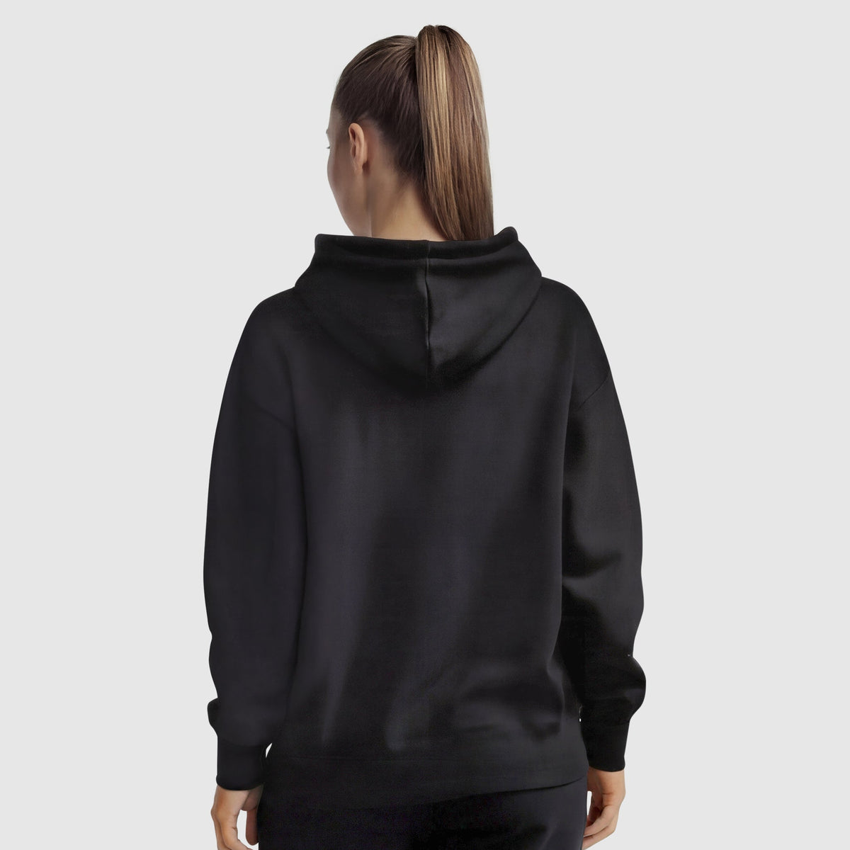 A woman with a high ponytail stands with her back to the camera, wearing a black Champion Powerblend Freshman Hoodie, C Logo, and black pants against a light gray background.