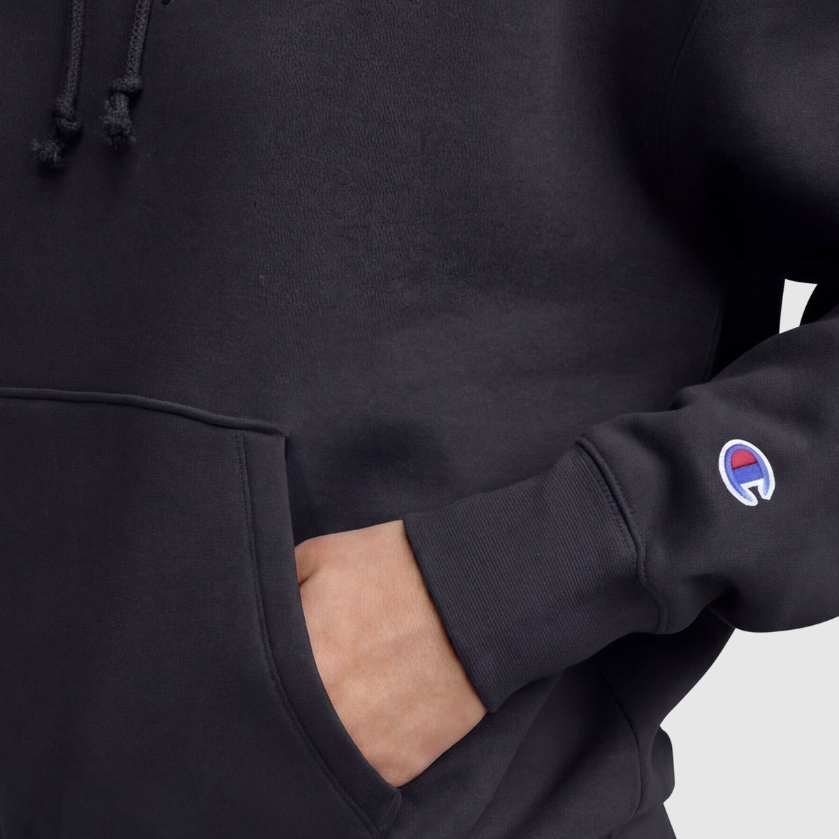 Close-up of a person wearing the Champion Powerblend Freshman Hoodie, C Logo, in black. One hand is in the front pocket, and the red, white, and blue C logo is embroidered on the sleeve, showcasing its cozy fleece fabric.