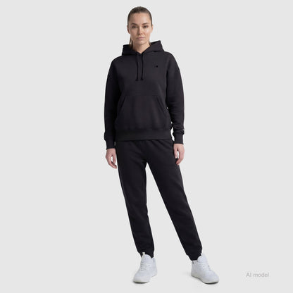 A person faces forward on a light gray background, wearing a black Champion Powerblend Freshman Hoodie with C Logo and matching joggers, one hand in their pocket. White sneakers complete the look, while the fleece adds extra comfort.