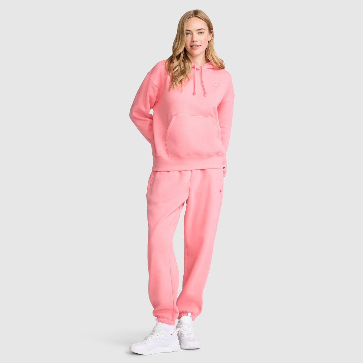 A woman stands wearing a pink Champion Small C Freshman Powerblend Hoodie and matching sweatpants with white sneakers. She has long blonde hair and is smiling slightly against a light gray background.