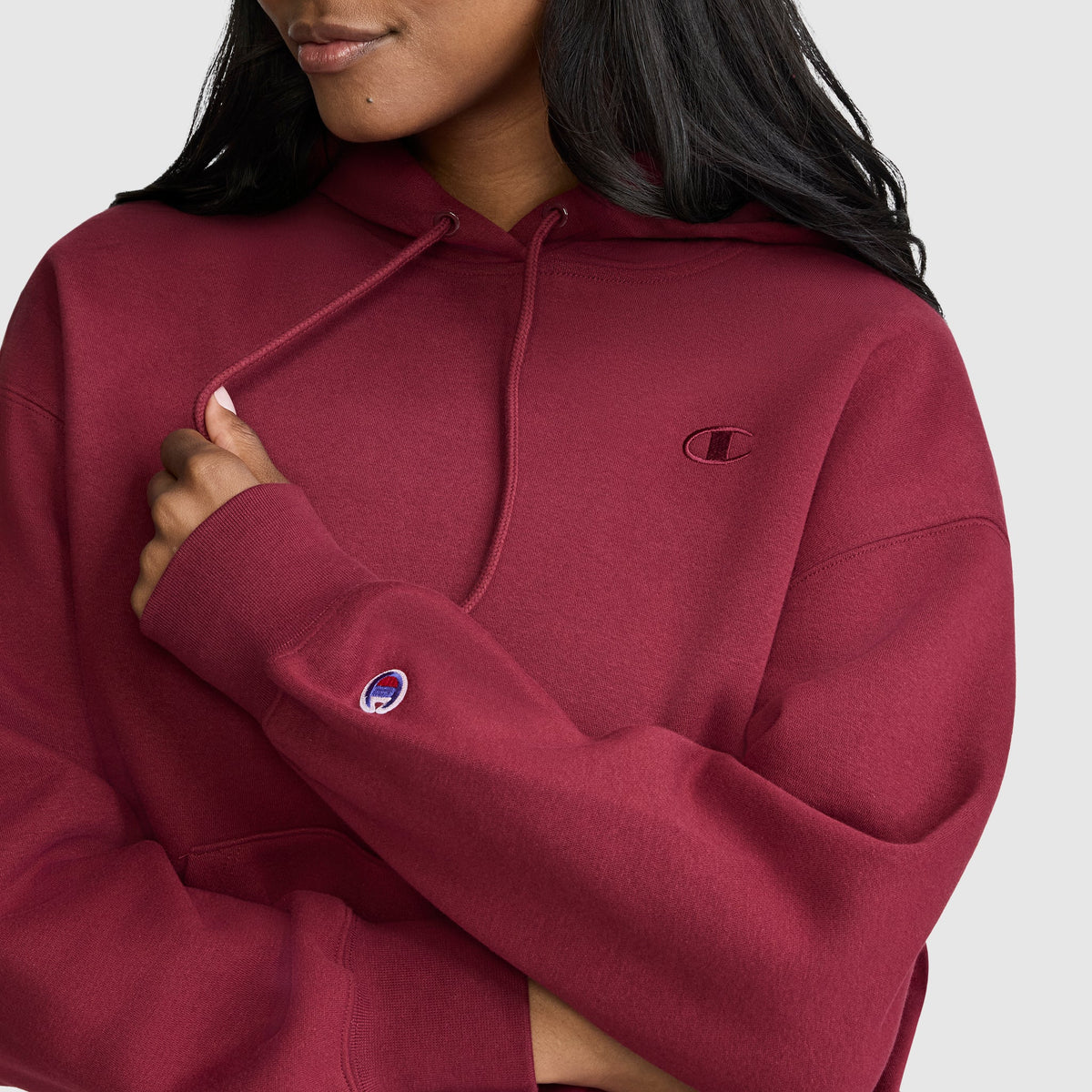 A woman wearing a maroon Champion Small C Freshman Powerblend Hoodie with the logo on the chest and sleeve stands with her arms crossed against a light gray background, highlighting the classic style and cozy comfort of this fleece women’s hooded sweatshirt.