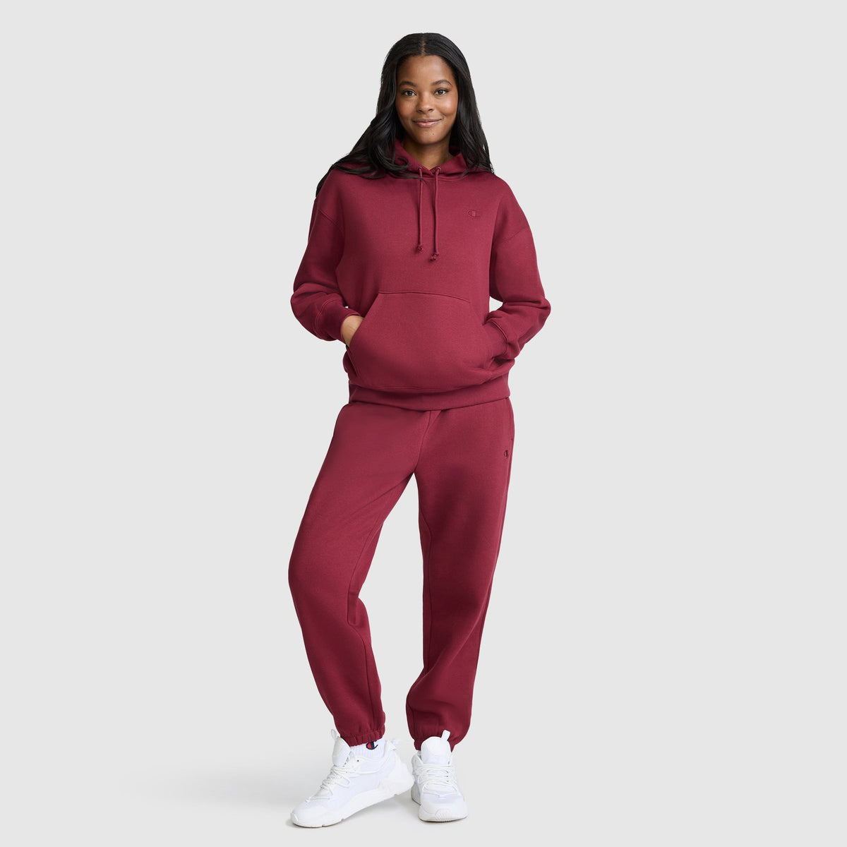 A person stands smiling against a plain light background, wearing the Champion Small C Freshman Powerblend Hoodie in maroon with matching sweatpants and white sneakers.