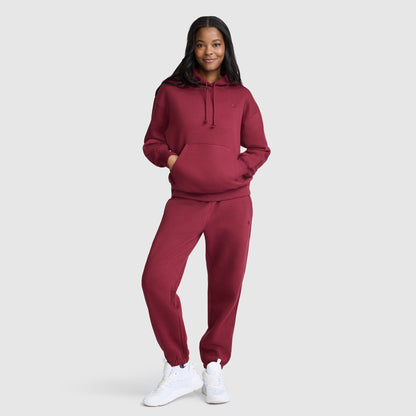 A person stands smiling against a plain light background, wearing the Champion Small C Freshman Powerblend Hoodie in maroon with matching sweatpants and white sneakers.