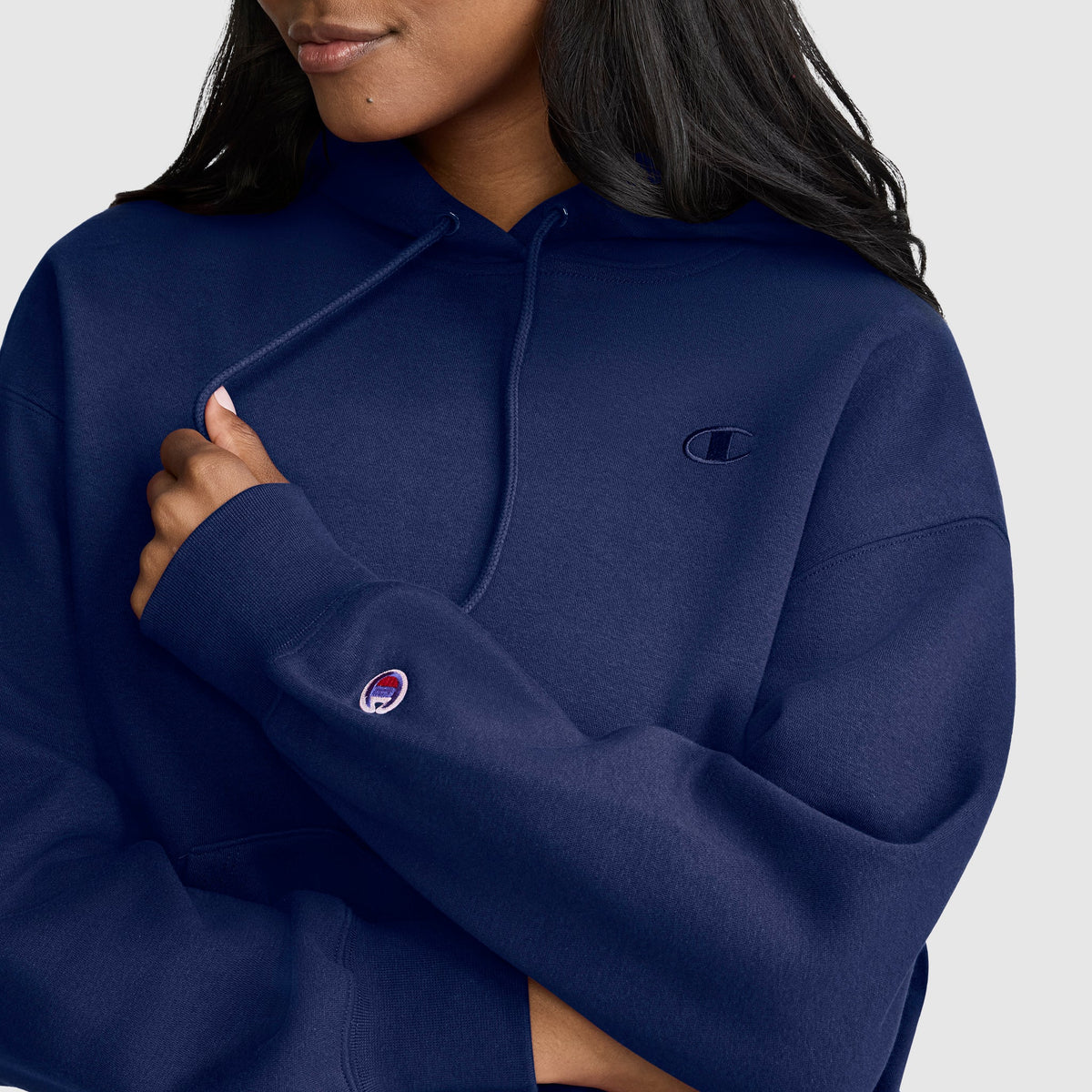 A woman with long dark hair is seen wearing the Champion Small C Freshman Powerblend Hoodie in navy blue, holding the drawstring in one hand. Only the lower half of her face is visible, and the classic Champion logo appears on both the chest and sleeve of this cozy women’s hoodie.